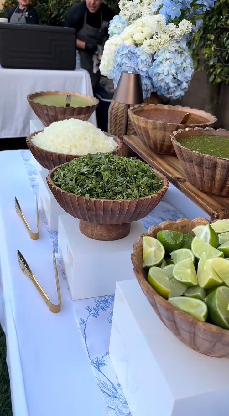 Lime wedges, cilantro, onions, and salsas in wooden bowls on a white buffet table.