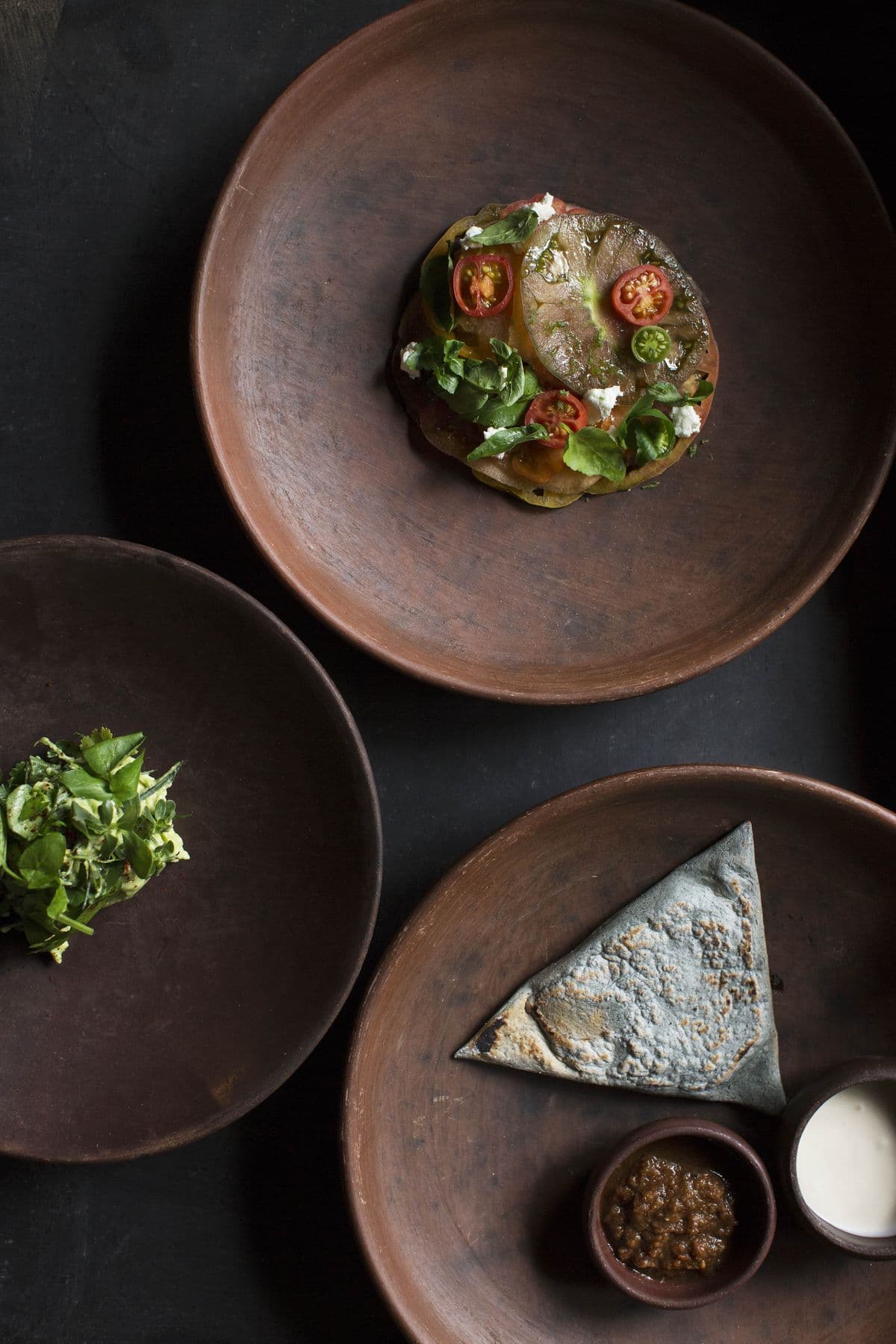 Three clay plates holding heirloom tomato salad, fresh greens, and a blue corn tortilla.