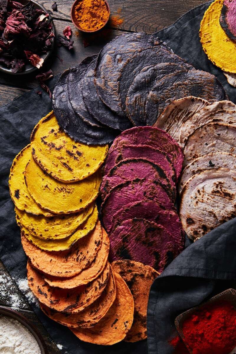 Colorful stacks of yellow, purple, orange, and blue corn tortillas arranged on dark fabric.
