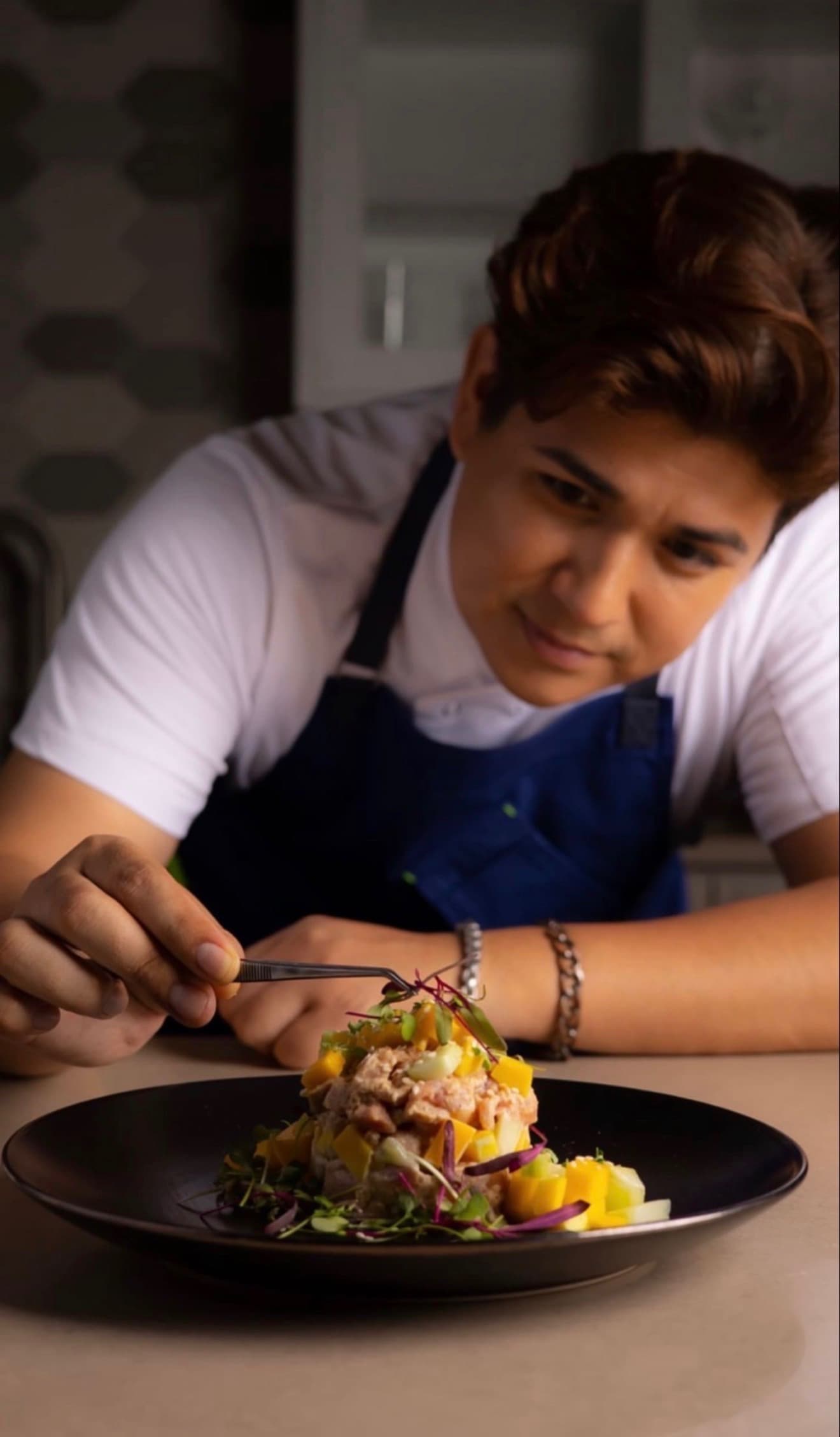 Focused chef uses tweezers to meticulously garnish a colorful seafood dish with fresh microgreens.