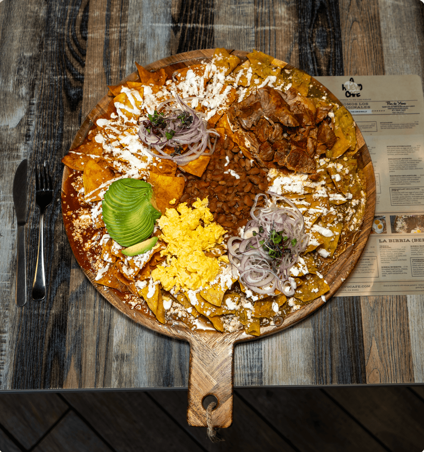 Large wooden platter of red and green chilaquiles with eggs, avocado, beans, and meat.