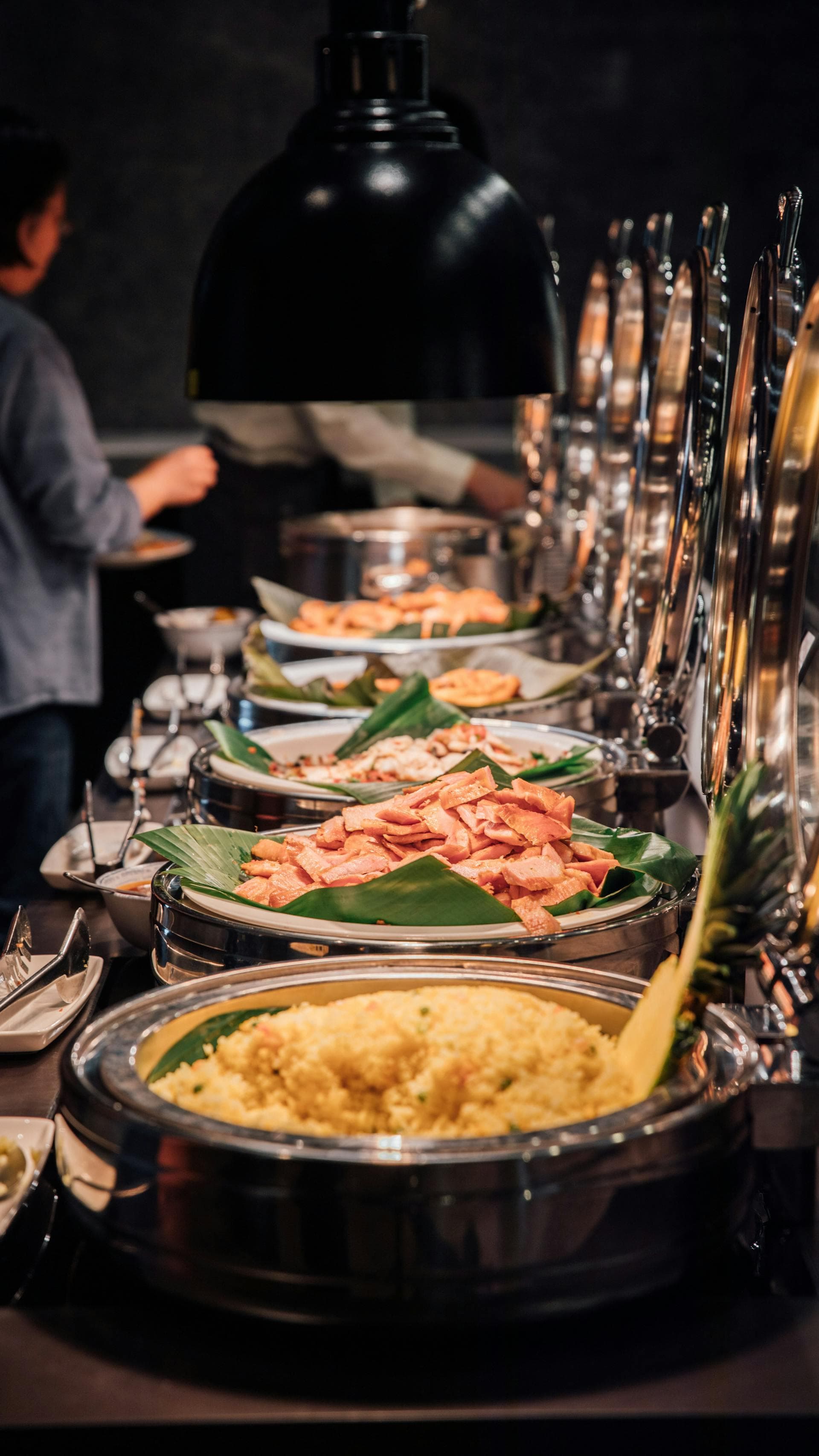 Silver buffet dishes containing yellow rice and sliced meat served on green banana leaves.