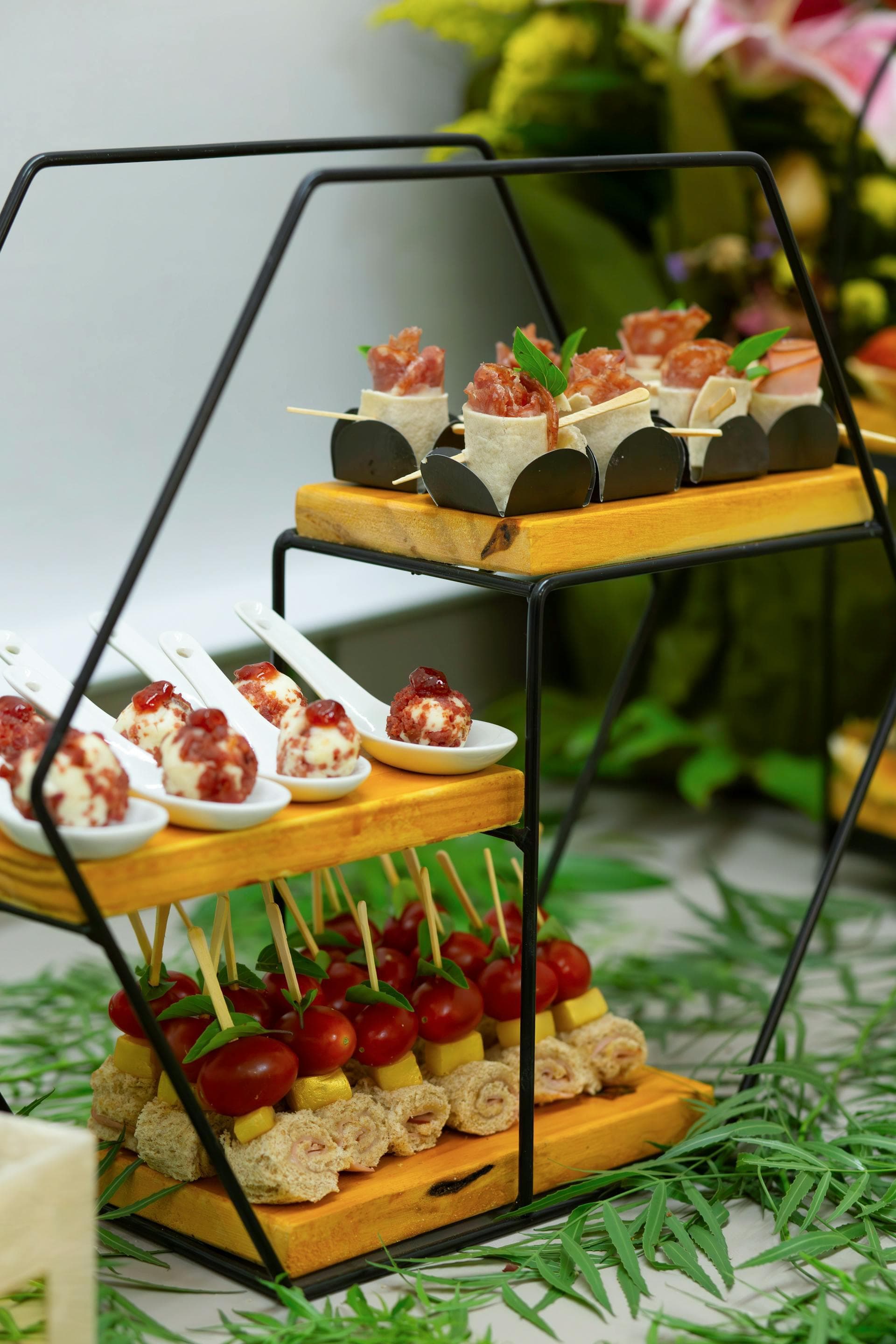 Three-tiered black stand displaying gourmet appetizers like meat wraps, cheese balls, and tomato skewers.