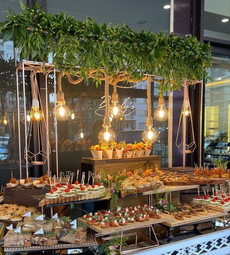 Elegant catering display with various appetizers under hanging light bulbs and lush green foliage.