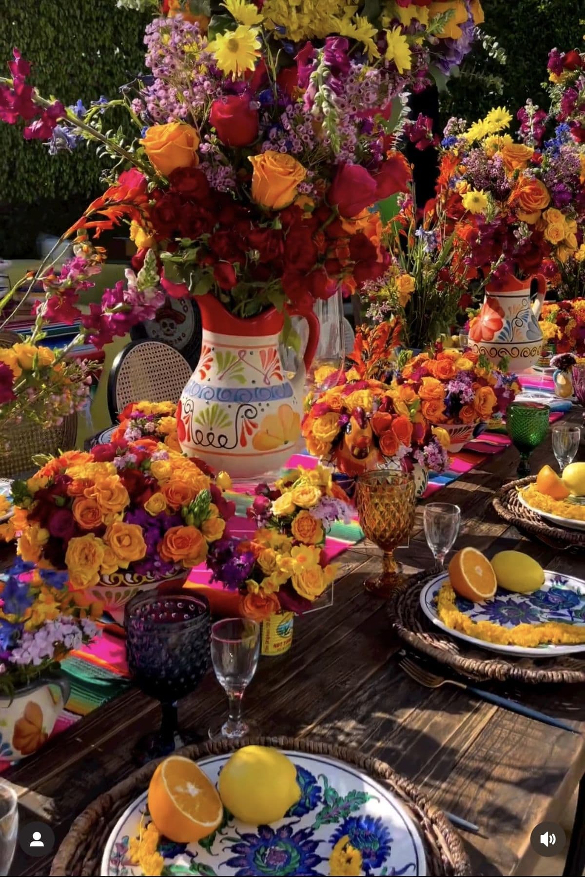 Vibrant outdoor table featuring colorful floral bouquets, painted pitchers, citrus fruits, and patterned dinnerware.