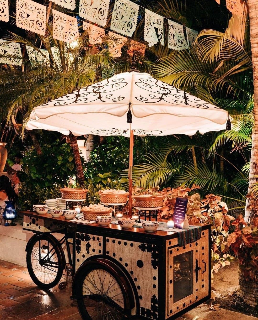 Decorative food cart with a patterned umbrella, tropical plants, and festive papel picado banners.